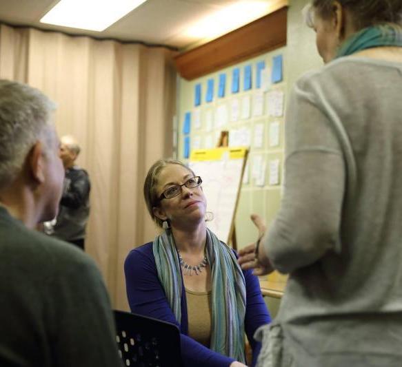 A group of adults gathered indoors, with a seated woman wearing glasses and a blue top and scarf looking toward two people standing nearby. A presentation board with notes and colored cards is visible in the background.
